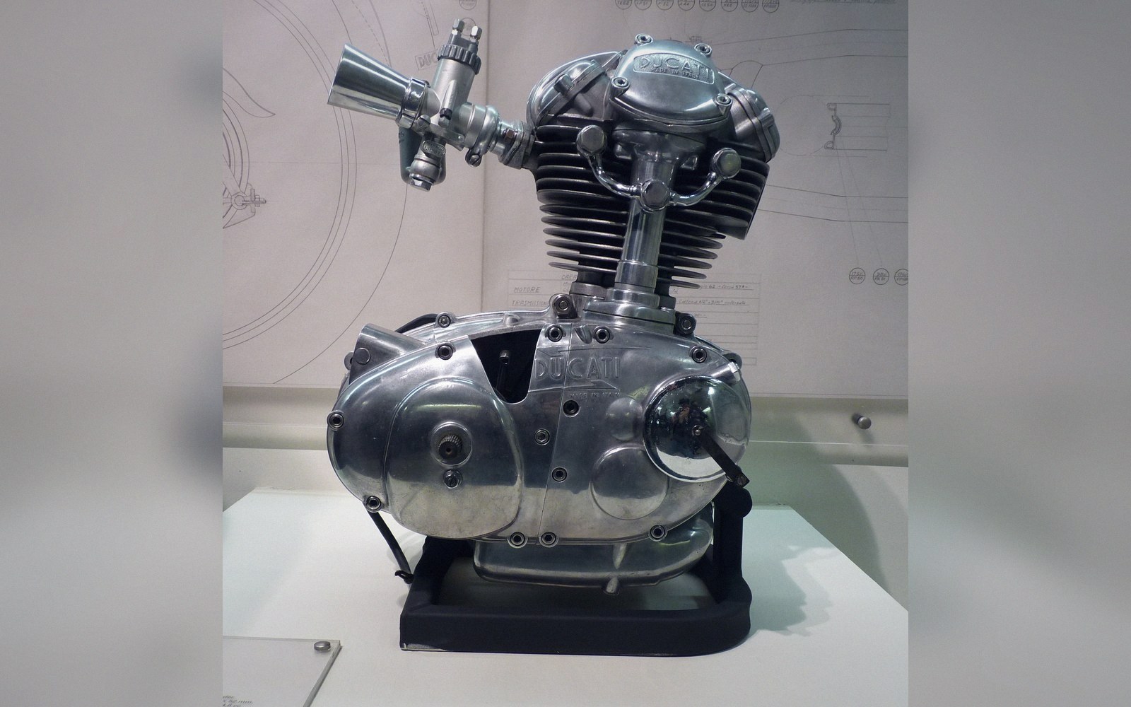 A single-cylinder Ducati engine displayed on a stand against technical blueprint drawings